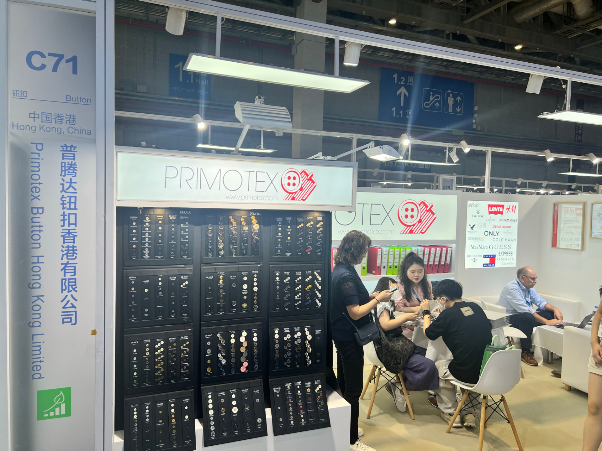 Wider view of two Primotex booth sections with customers and visitors, customer logos displayed in the background