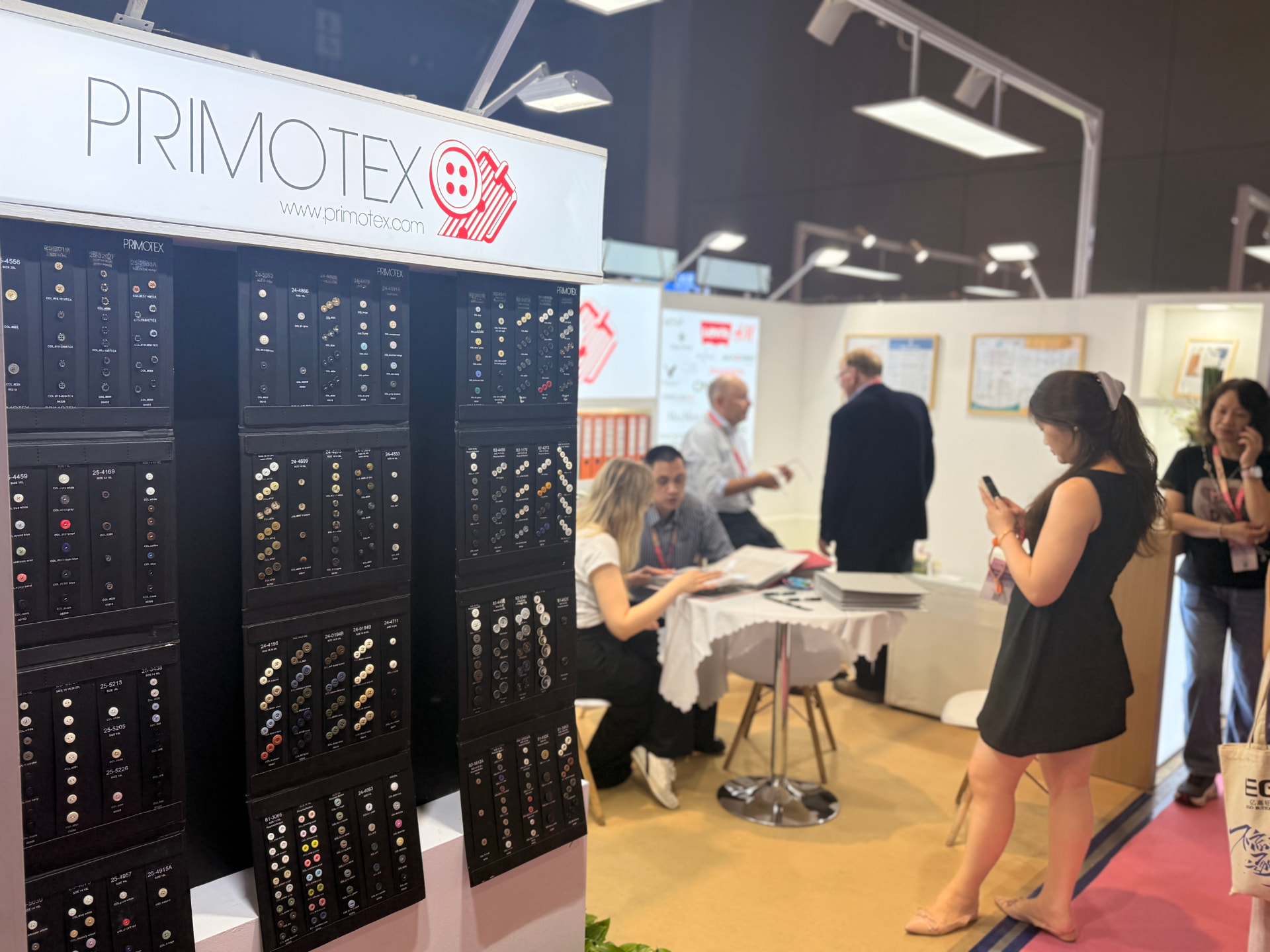 Primotex button sample wall in the foreground with visitors meeting at a table inside the booth
